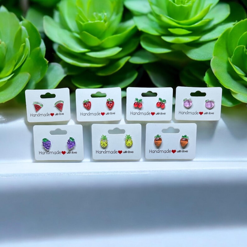 以下が含まれることがあります： 「Handmade with love」というテキストが書かれた白いカードに、フルーツをモチーフにしたスタッドピアスの詰め合わせ。ピアスは、スイカ、イチゴ、チェリー、パイナップル、ニンジン、ブドウをモチーフにしている。