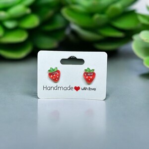 以下が含まれることがあります： 白いカードに「Handmade with love」と書かれた、赤いイチゴのスタッドピアスのペアです。