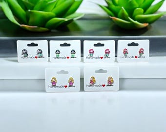 May include: Six pairs of small, colourful, cartoon character earrings on white cards with the text "Handmade with love".