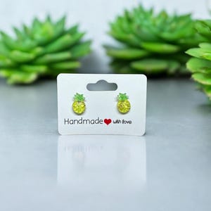 以下が含まれることがあります： 白いカードに「Handmade with love」と書かれた、黄色のパイナップルスタッドイヤリングのペアです。