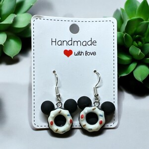 May include: A pair of silver dangle earrings with black and white donut charms shaped like Mickey Mouse ears. The earrings are on a white card with the text "Handmade with love".