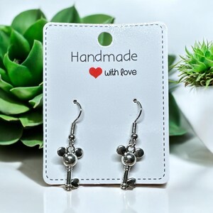 May include: Silver key-shaped earrings with a Mickey Mouse head charm. The earrings are on a white card with the text "Handmade with love".