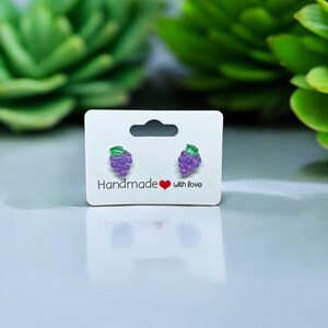 以下が含まれることがあります： 白いカードに「Handmade with love」と書かれた、紫色のブドウのスタッドピアスのペアです。