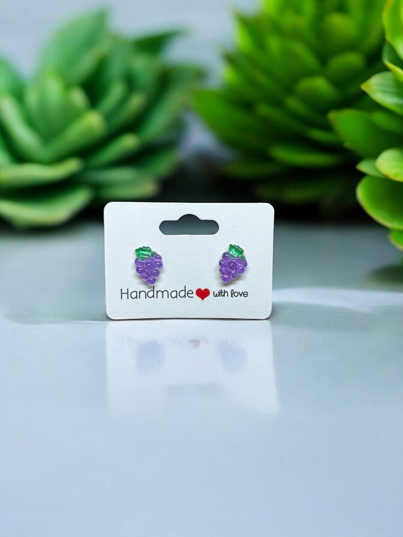 以下が含まれることがあります： 白いカードに「Handmade with love」と書かれた、紫色のブドウのスタッドピアスのペアです。