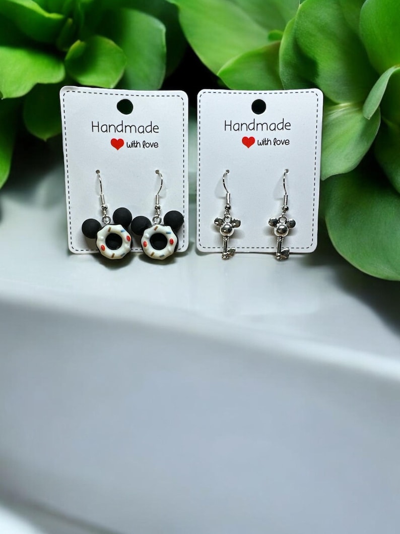 May include: Two pairs of dangle earrings displayed on white cards. The first pair features a black Mickey Mouse head with a white and red donut. The second pair features a silver key with a Mickey Mouse head.
