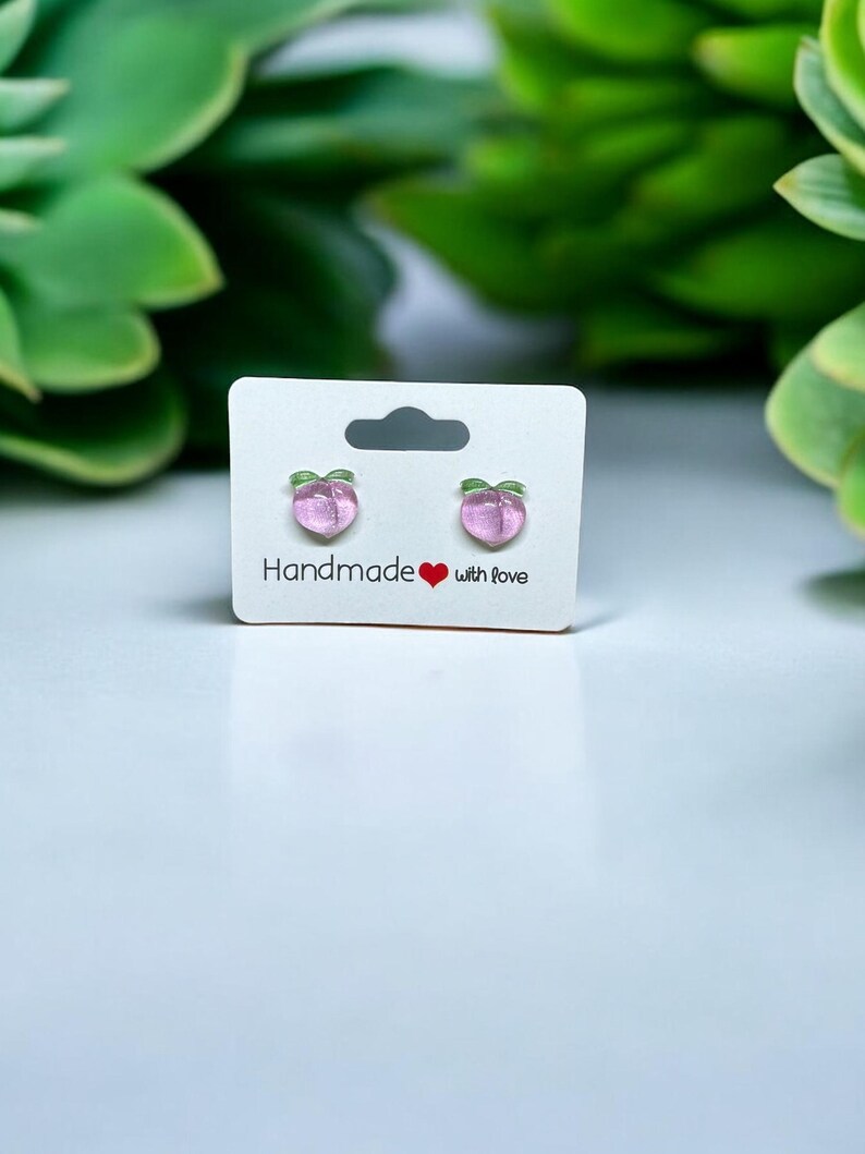 以下が含まれることがあります： 白いカードに「Handmade with love」と書かれた、桃の形をしたイヤリングのペアです。