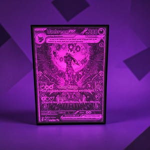 May include: A framed, illuminated artwork featuring a Pokémon trading card. The card, with a purple and black design, displays the text "Umbreon EX" and "Moon Mirage." The artwork is encased in a black frame, and the background is a gradient of purple.