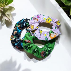 Puede incluir: Tres scrunchies de pelo coloridos con temática de Minecraft. Los scrunchies son verdes, rosas y negros con diferentes personajes y bloques de Minecraft impresos en ellos.