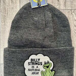 May include: Gray knit beanie with a white cloud-shaped patch featuring the text "BILLY STRINGS IS A NATURAL HIGH" and a green frog illustration. A blue tag hangs from the beanie.