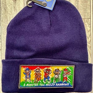 May include: A navy blue beanie with a rectangular patch featuring cartoon characters playing instruments. The patch reads "5 MINUTES TILL FUZZY RAINBOWS" in white text. A tag with the word "GELANTE" is attached.