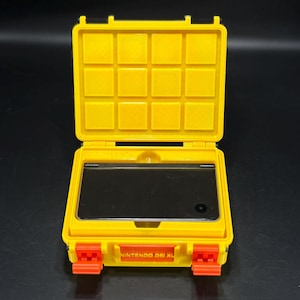 Puede incluir: Estuche protector amarillo para una Nintendo DSi XL, abierto para revelar la consola. El estuche tiene cierres naranjas y el texto "NINTENDO DSi XL" es visible. El estuche está diseñado para proteger la consola.
