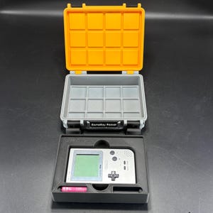 May include: An open, yellow and gray Game Boy Pocket case with a silver Game Boy Pocket console inside. The case is open, revealing a gray interior with a grid pattern. A pink battery is visible. The case is labeled "GameBoy Pocket".