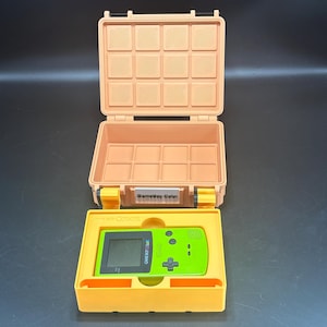 Puede incluir: Un estuche melocotón abierto, con un patrón de rejilla en la tapa y el interior. Dentro, una consola Game Boy Color verde brillante descansa en una bandeja amarilla. El texto "Game Boy Color" es visible en el estuche y la consola.
