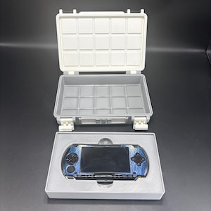 Puede incluir: Un maletín de transporte de plástico gris y blanco, abierto para revelar un interior gris. Debajo, una bandeja gris contiene una consola de juegos portátil PSP azul y negra. La funda tiene un asa y cierres. La PSP tiene un diseño azul y negro.