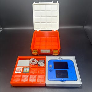 Puede incluir: Un estuche abierto naranja y blanco con asa, que contiene una consola Nintendo 2DS azul y cartuchos de juego. El estuche tiene una bandeja para la consola y los cartuchos de juego, y un compartimento separado para accesorios.