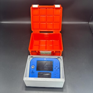 Puede incluir: Un estuche naranja brillante, abierto para revelar un interior cuadriculado, se encuentra encima de una consola Nintendo 2DS azul en una caja blanca. La consola tiene una pantalla y botones, con "NINTENDO 2DS" impreso en la caja.