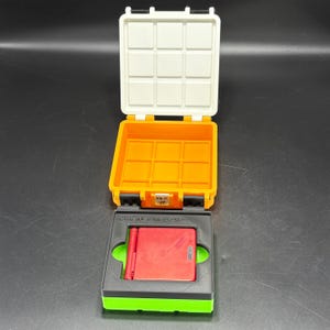 Puede incluir: Un estuche de plástico naranja y blanco abierto con un dispositivo de juego portátil rojo en el interior. El estuche es rectangular con una tapa con bisagras y un pestillo negro. El dispositivo portátil es rojo con un botón verde. El estuche está sobre una base negra y verde.