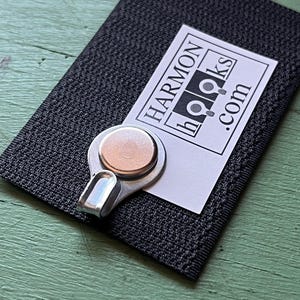 May include: A black fabric-covered hook with a silver metal hook and a round magnet. The hook has a white label with the text "HARMON HOOKS .com".