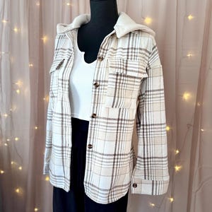 May include: A cream and brown plaid shirt jacket with a hood, displayed on a mannequin. The jacket has a button-up front, two chest pockets, and a white undershirt. The mannequin is wearing black trousers.