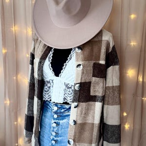 Puede incluir: Un sombrero fedora beige está sobre un maniquí que lleva una camisa abotonada a cuadros marrón y beige, una blusa de encaje blanca y unos vaqueros azules de cintura alta. La camisa tiene botones grandes. El fondo es de color neutro.