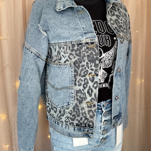 May include: A blue denim jacket with a leopard print panel on the front. The jacket has a button closure and two pockets on the front.