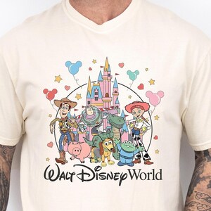 Puede incluir: Camiseta de color marfil con un gr&aacute;fico colorido de los personajes de Toy Story de Disney frente a un castillo, con la inscripci&oacute;n "Walt Disney World". La camiseta tiene cuello redondo y mangas cortas.