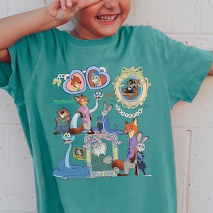 May include: A teal-colored t-shirt featuring a colorful cartoon graphic of characters from Zootopia. The graphic includes images of Nick Wilde, Judy Hopps, and other characters. The shirt is worn by a young girl.