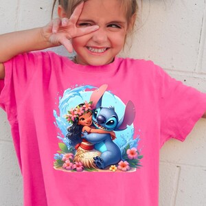 May include: A pink t-shirt with a graphic of Stitch from Lilo & Stitch hugging Moana from Moana. The graphic is set against a blue wave background with pink flowers.