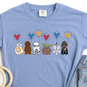 May include: A light blue t-shirt with a graphic of Star Wars characters holding Mickey Mouse balloons. The characters include Chewbacca, a Stormtrooper, Yoda, R2-D2, and Darth Vader.