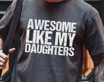 May include: A charcoal gray short-sleeve t-shirt featuring the white distressed-style text "AWESOME LIKE MY DAUGHTERS" printed across the chest. The shirt has a crew neckline and a relaxed fit, showcasing a weathered fabric appearance.