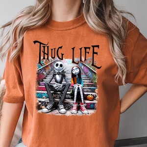 Puede incluir: Camiseta naranja con un estampado gr&aacute;fico de Jack Skellington y Sally de Pesadilla Antes de Navidad sentados en un tramo de escaleras con grafitis. El texto "THUG LIFE" est&aacute; impreso sobre los personajes.