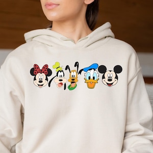 Op de afbeelding: Een cr&egrave;mekleurige hoodie met een afbeelding van Mickey Mouse, Minnie Mouse, Donald Duck, Goofy en Pluto. De personages staan in een rij, met hun gezichten naar voren gericht. De hoodie heeft een capuchon met trekkoord en een kangoeroezak.