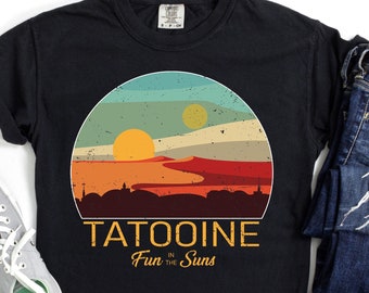 May include: Black t-shirt with a retro sunset graphic featuring two suns over a desert landscape. The text "TATOOINE Fun in the Suns" is printed below the graphic. The shirt is paired with blue jeans and gray sneakers.