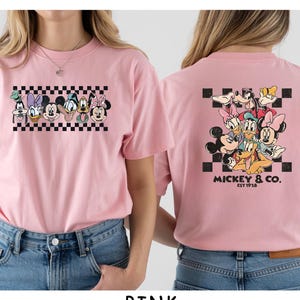 May include: A light pink t-shirt featuring a graphic of Disney characters on the front and back. The front shows a horizontal row of character faces over a black and white checkerboard pattern. The back displays a larger group of characters.