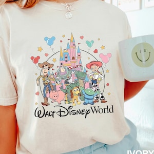 Puede incluir: Camiseta color marfil con un gr&aacute;fico colorido de los personajes de Toy Story frente a un castillo, con las palabras "Walt Disney World" debajo. Tambi&eacute;n se ve una taza blanca con una carita sonriente.