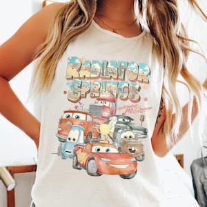 Puede incluir: Camiseta sin mangas color marfil con un gr&aacute;fico colorido de personajes de la pel&iacute;cula Cars, incluido Rayo McQueen. El texto "Radiator Springs" se muestra encima de los personajes en una fuente retro.
