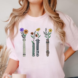 Puede incluir: Camiseta rosa con un gr&aacute;fico de cuatro sables de luz con flores en los mangos. Los sables de luz son verde, azul, morado y rojo.