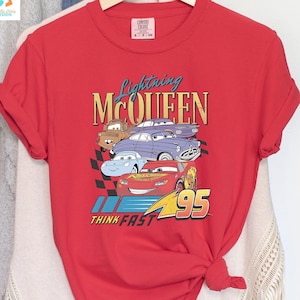 May include: Red t-shirt featuring a graphic of Lightning McQueen and other Cars characters. The text "Lightning McQueen" and "Think Fast" are printed on the shirt.