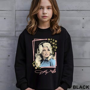 May include: A black long-sleeve sweatshirt featuring a graphic print of a blonde person with voluminous hair wearing a blue denim shirt. The design includes a pink rectangular frame, leopard print accents, a butterfly, and the script text Dolly.