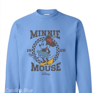Puede incluir: Una sudadera de color azul claro con un gr&aacute;fico de estilo vintage de Minnie Mouse. El gr&aacute;fico presenta a Minnie Mouse con un vestido azul y zapatos rojos, con el texto "Minnie Mouse" y "1928" en un c&iacute;rculo a cuadros blanco y negro. El texto "Disney" tambi&eacute;n est&aacute; incluido en el gr&aacute;fico.