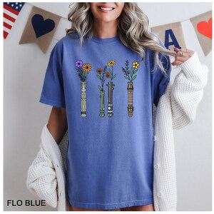 Puede incluir: Una camiseta azul con un gr&aacute;fico de cuatro sables de luz con flores que crecen desde la parte superior. Los sables de luz son verde, azul, rojo y morado.