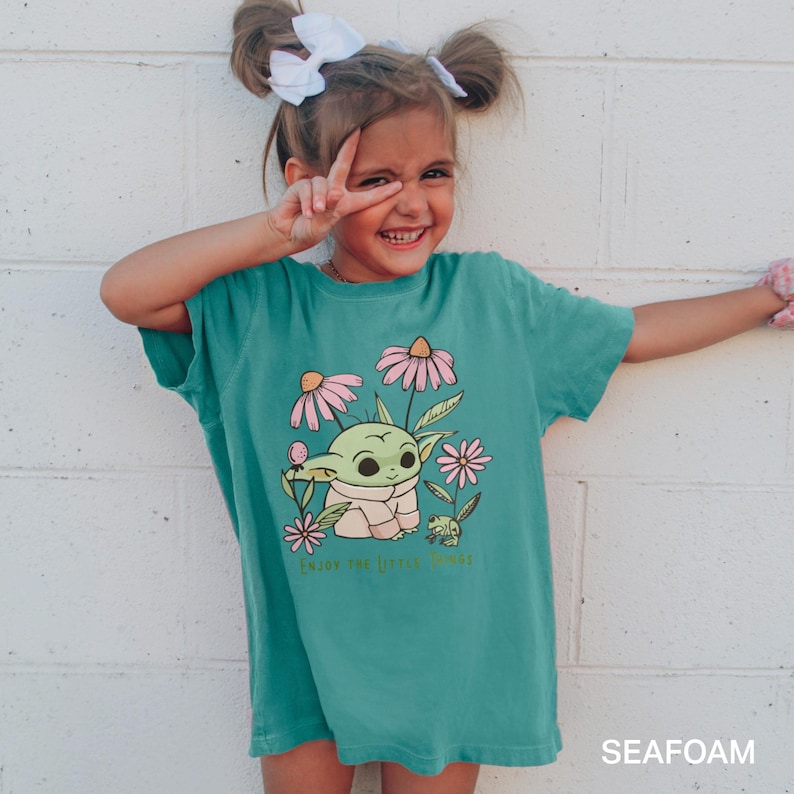Puede incluir: Una camiseta azul turquesa con una imagen de dibujos animados de un personaje verde con ojos grandes y flores rosas. El texto "Enjoy the little things" est&aacute; impreso en la camiseta.