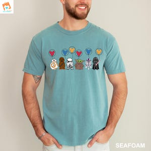 May include: A teal blue t-shirt with a graphic of Star Wars characters holding Mickey Mouse-shaped balloons. The characters include Chewbacca, Darth Vader, R2-D2, Yoda, and a Stormtrooper.