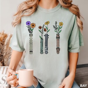 Puede incluir: Una camiseta azul claro con un gr&aacute;fico de cuatro sables de luz con flores en la parte superior de cada sable de luz. Las flores son moradas, naranjas, amarillas y rojas.