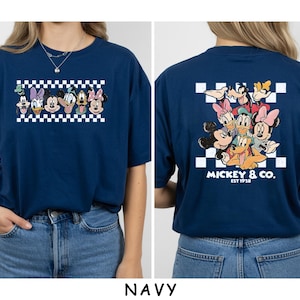 May include: A navy blue t-shirt featuring a front graphic of Disney characters in a white and navy checkered rectangle. The back displays a larger group of Disney characters over a checkered square with the text MICKEY & CO. EST 1928.