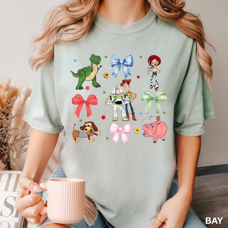 May include: A light green t-shirt with a variety of Toy Story characters and bows printed on it. The characters include Woody, Buzz Lightyear, Rex, Slinky Dog, Hamm, and Jessie. The bows are in various colors, including pink, green, and blue.