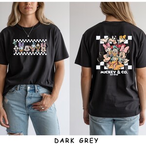 May include: A dark grey t-shirt featuring a front graphic of Disney characters in a black and white checkered box. The back displays a larger version of the same characters with the text MICKEY & CO. EST 1928 printed in white below the design.