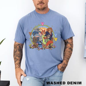 Op de afbeelding: Een lichtblauw t-shirt met een kleurrijke cartoonillustratie van verschillende dierenfiguren. De afbeelding bevat een vos, een leeuw, een konijn en andere dieren, omringd door bloemen en ballonnen. De tekst "WASHED DENIM" staat onderaan het shirt.