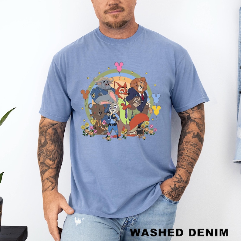 Op de afbeelding: Een lichtblauw t-shirt met een kleurrijke cartoonillustratie van verschillende dierenfiguren. De afbeelding bevat een vos, een leeuw, een konijn en andere dieren, omringd door bloemen en ballonnen. De tekst "WASHED DENIM" staat onderaan het shirt.