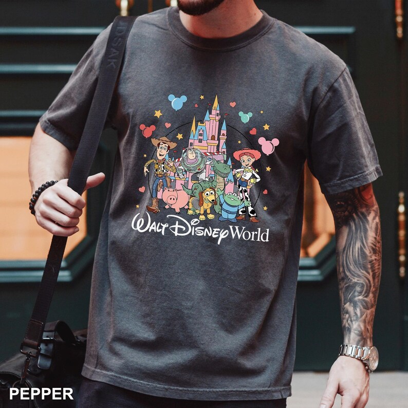 Puede incluir: Camiseta gris oscuro con un gr&aacute;fico colorido de personajes de Disney, incluyendo a Woody, Buzz Lightyear y Jessie, frente a un castillo. Las palabras "Walt Disney World" est&aacute;n impresas debajo de los personajes.